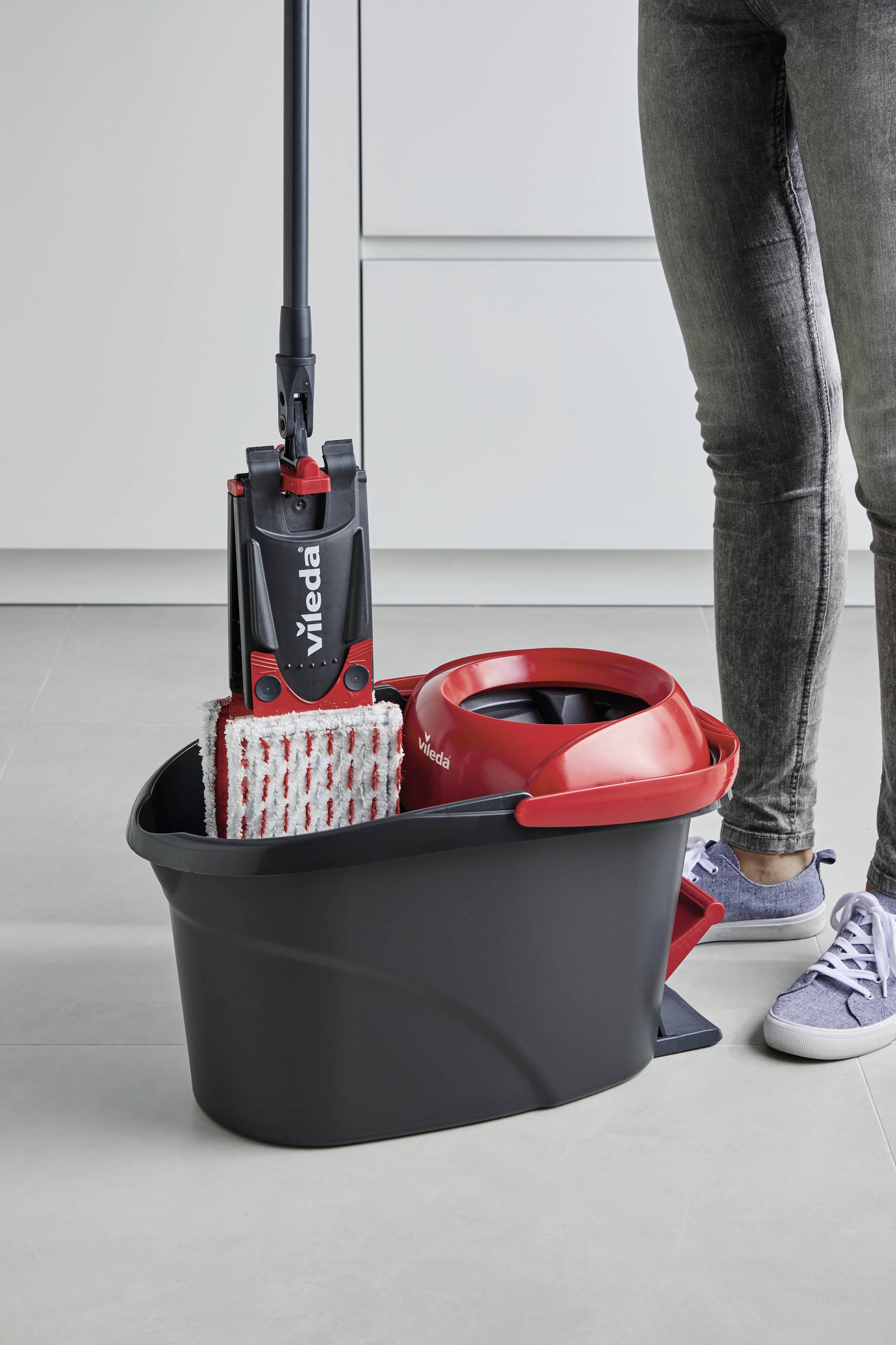 Wischmoppset mit rotem Eimer und schwarzem Stiel wird von einer Person mit Jeans und Sneakern bedient auf einem Küchenfliesenboden.