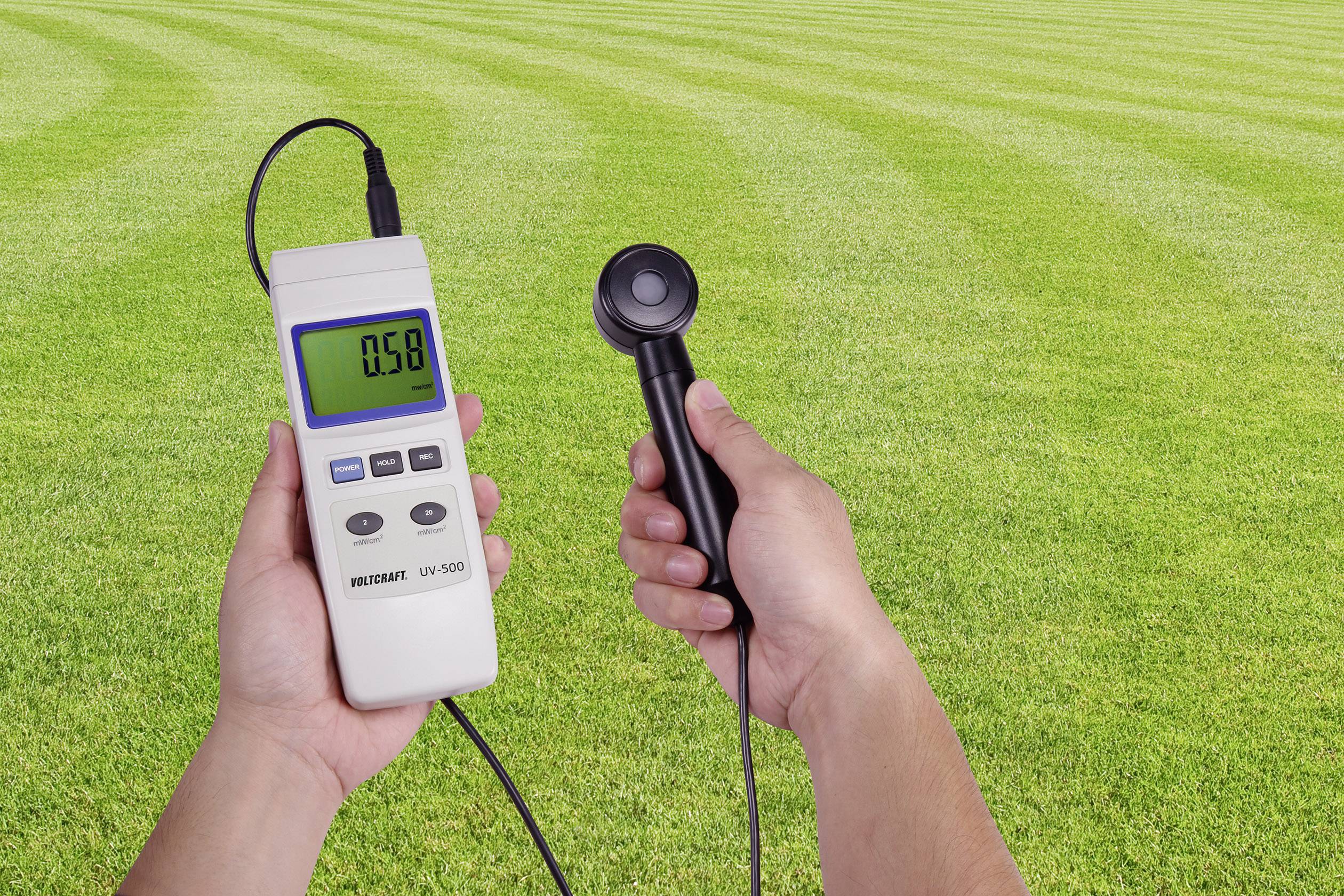 Eine Person hält ein UV-Messgerät über einer grünen Rasenfläche. Das Display zeigt den Wert '0,59' an.