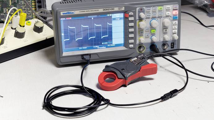 Auf einem Tisch steht ein Oszilloskop mit einem Farbdisplay und einem Bedienfeld mit mehreren Reglern. Daran angeschlossen ist ein rot-schwarzer Stromzangenadapter. Ein Großteil des Verbindungskabels ist eingerollt und mit einem Kabelbinder fixiert. Im Hintergrund befindet sich eine weitere Bedienstation mit mehreren Drehreglern.