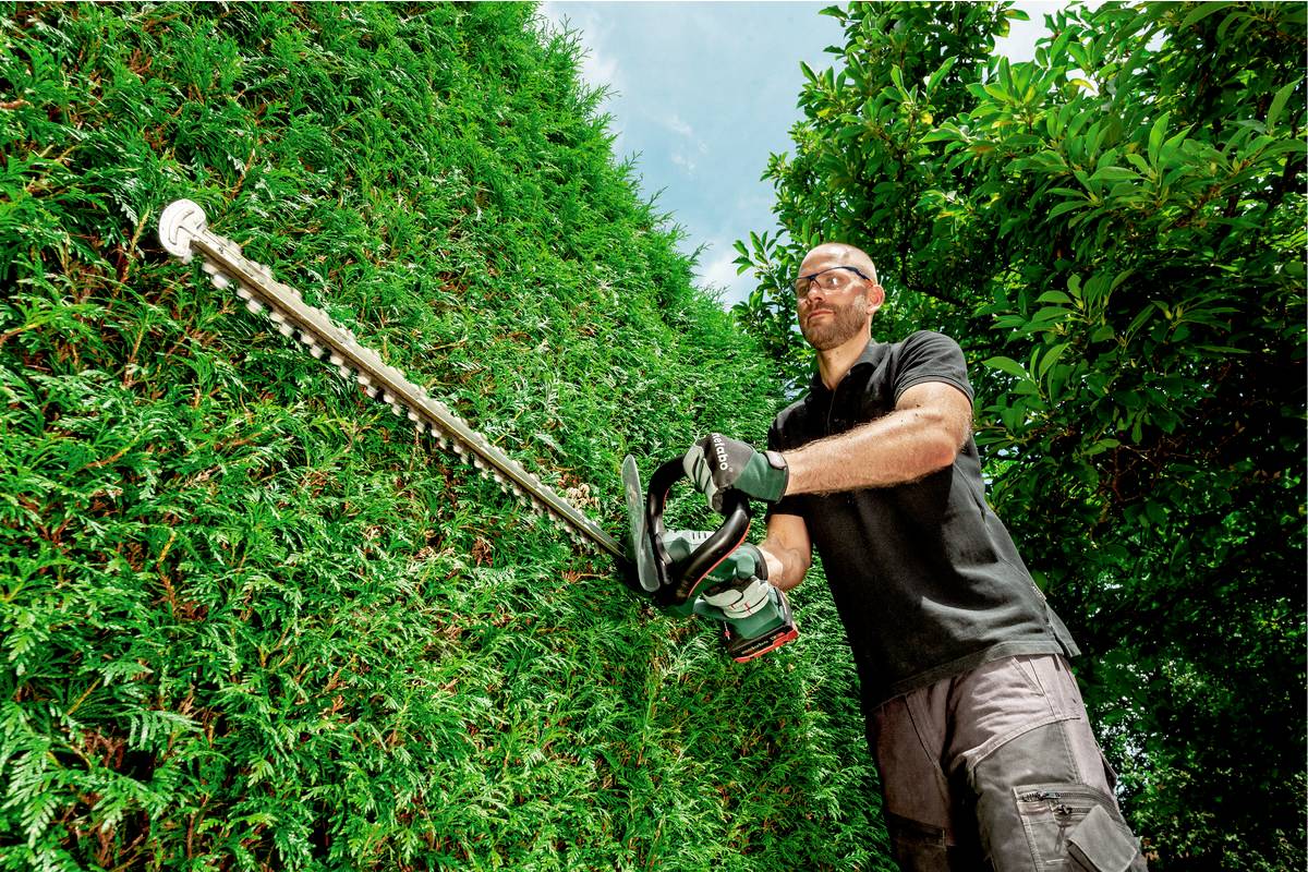 Ein Mann schneidet eine hohe grüne Hecke mit einer motorisierten Heckenschere. Er trägt Schutzbrille und Handschuhe unter klarem Himmel.