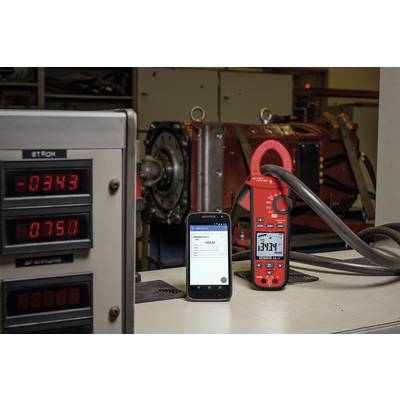Ein digitales Multimeter misst den Strom in einer Werkstatt. Ein Smartphone zeigt die gleiche Messung an. Im Hintergrund eine Maschine.