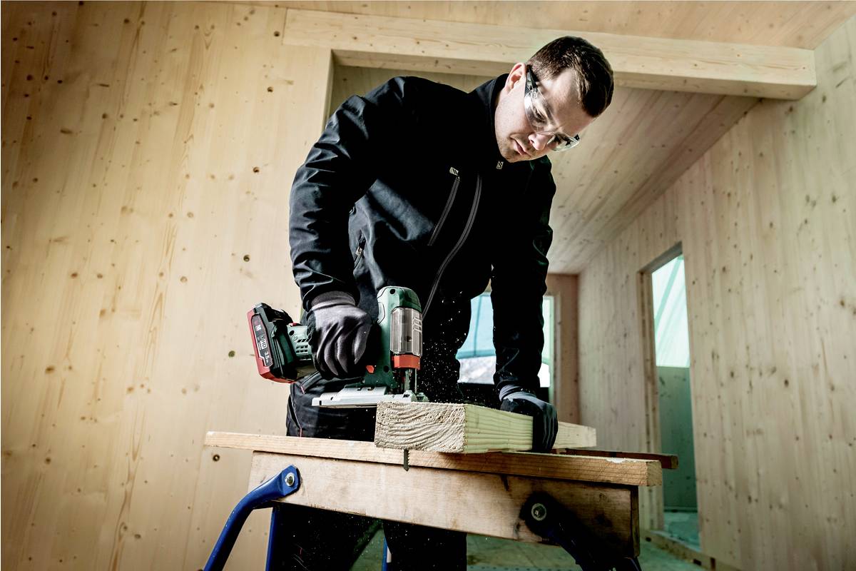 Ein Mann in Schutzbrille und Handschuhen bohrt mit einer Bohrmaschine in ein Holzstück auf einer Werkbank in einem Holzraum.