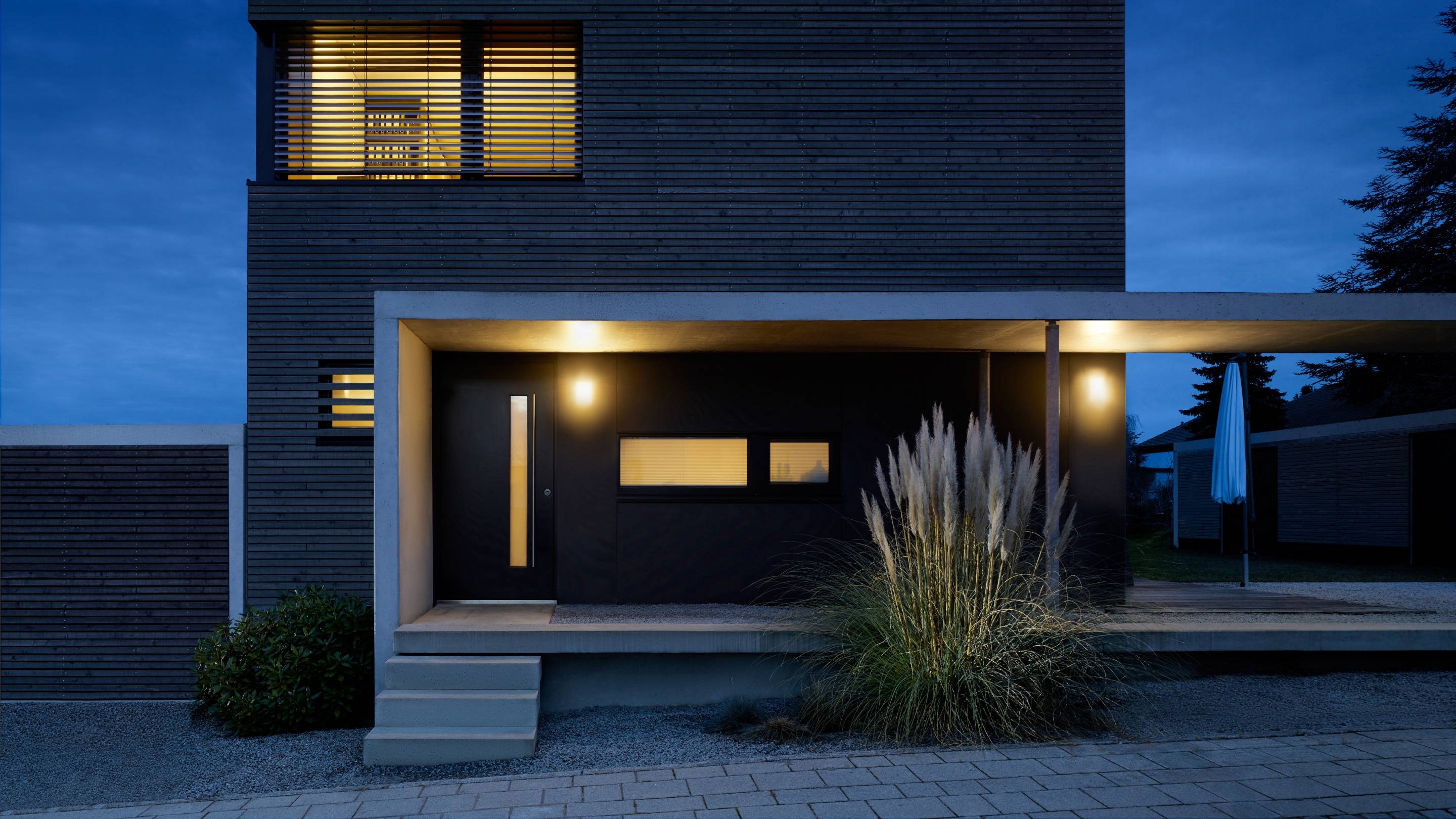 Modernes Haus bei Nacht mit leuchtenden Außenlichtern und großen Fenstern. Dunkle Architektur, umgeben von Pflanzen und einer ruhigen Umgebung.