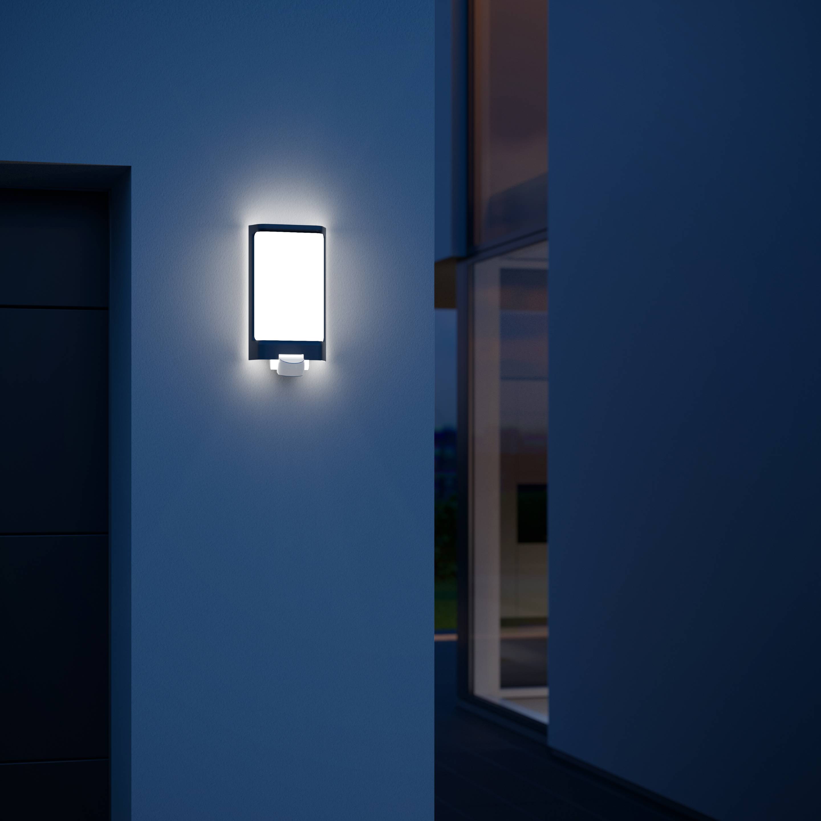 Eine moderne Außenlampe leuchtet an einer Wand eines Gebäudes in der Dämmerung. Die Umgebung ist dunkel und die Lampe strahlt hell.