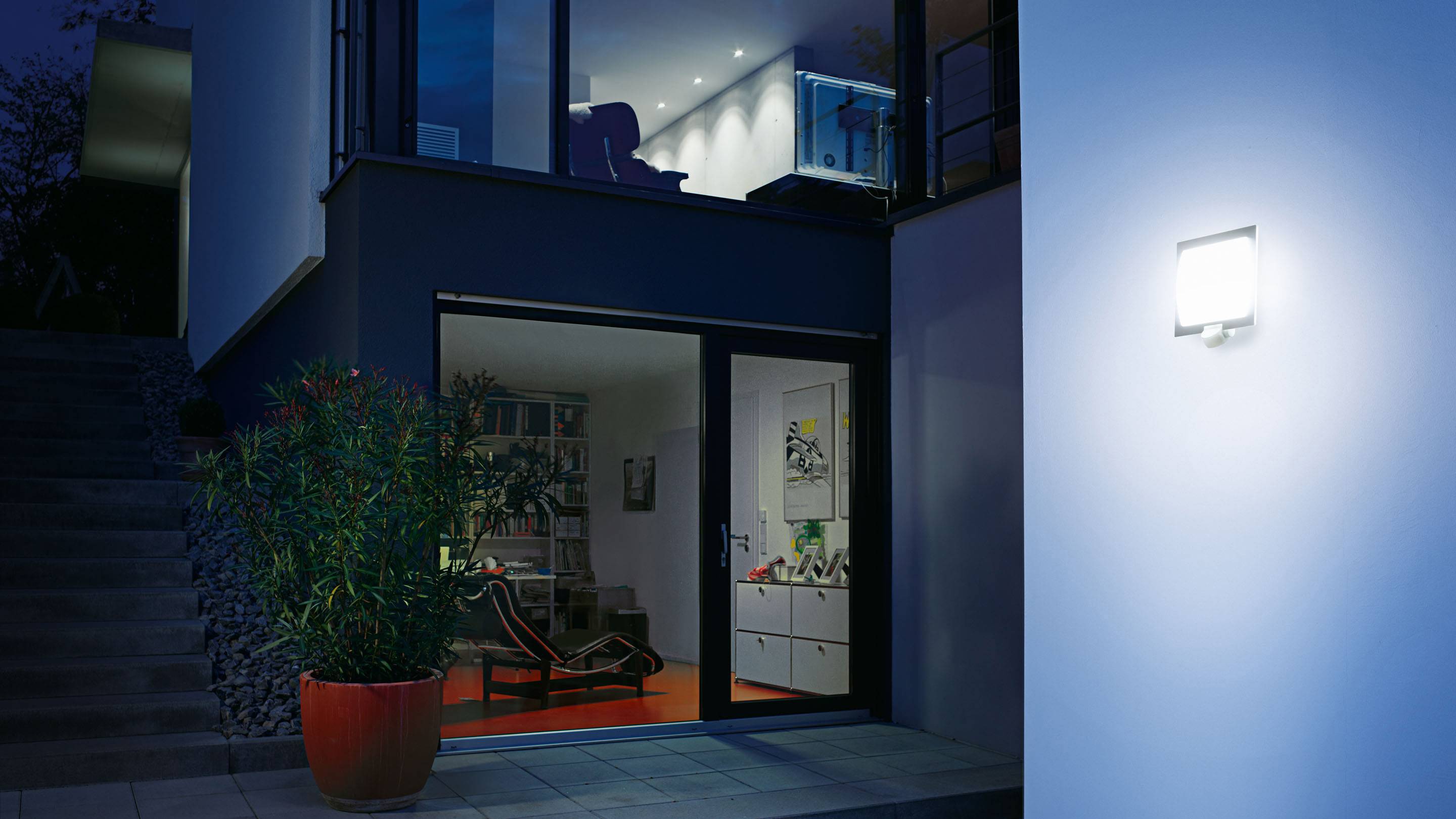 Ein modernes Haus bei Nacht mit eingeschaltetem Außenlicht, das den Eingangsbereich erleuchtet. Eine große Pflanze steht links neben der Tür.