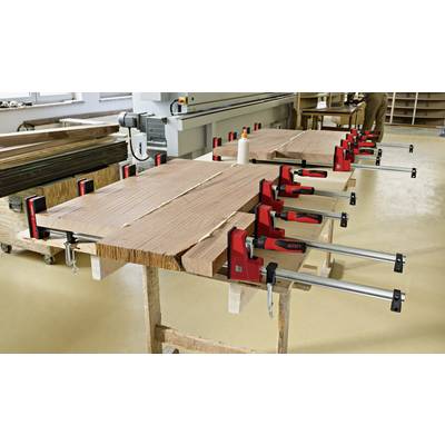 Mehrere Holzplatten werden mit roten Metallzwingen festgehalten, um sie zusammenzupressen. Im Hintergrund ist eine Werkstatt zu sehen.