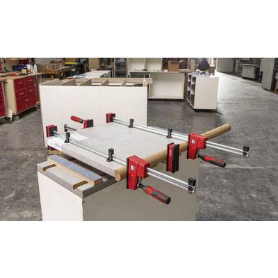 Eine Werkbank mit mehreren Klammern, die ein Holzstück fixieren. Im Hintergrund sind weitere Werkzeuge und Möbelstücke in einer Werkstatt sichtbar.