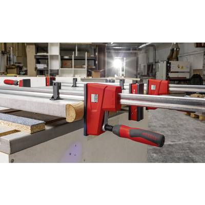 Mehrere rote Schraubzwingen fixieren Holzbretter auf einer Werkbank in einer Tischlerwerkstatt. Im Hintergrund sind Regale zu sehen.