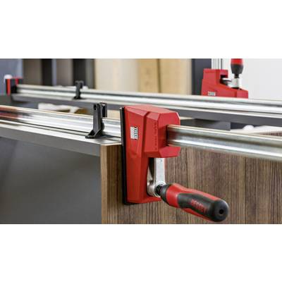Eine rote Schraubzwinge hält ein Holzstück fest. Die Klemme ist auf einer Metallstange befestigt, die Stabilität für Tischlerarbeiten bietet.