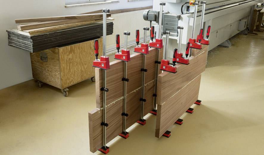 Holzplatten werden in einer Werkstatt mit großen Schraubzwingen zusammengepresst. Eine Maschine ist im Hintergrund sichtbar.