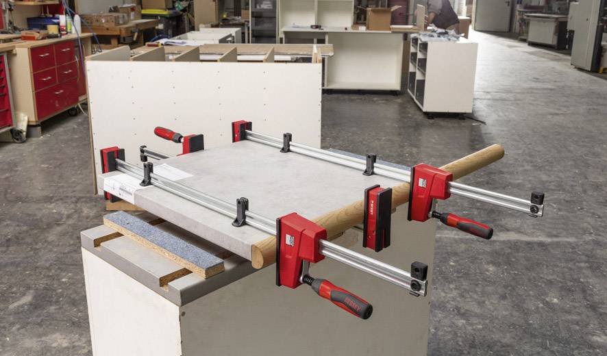 Ein Werkstück wird mit mehreren Zwingen auf einem Tisch in einer Werkstatt fixiert. Hintergrund zeigt Werkbank und Regale.