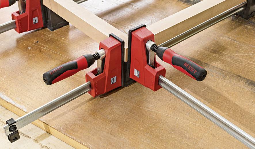 Zwei rote Schraubzwingen halten Holzbalken in einem rechten Winkel auf einem Tisch zusammen.