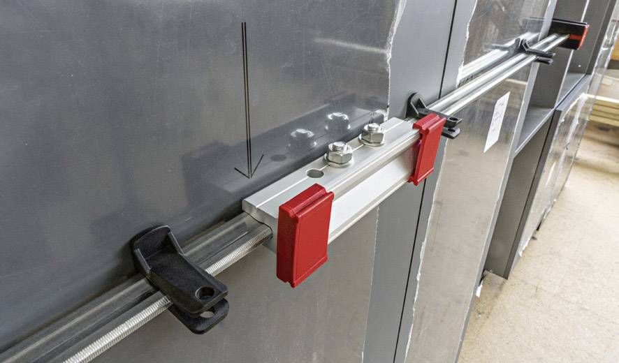 Eine Metallkonstruktion mit Schrauben und roten Klemmen befestigt eine Leiste an einer glatten, dunklen Oberfläche.