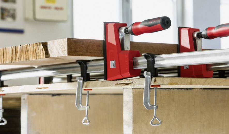 Zwei rote Schraubzwingen halten ein Holzbrett auf einem Tisch. Sie sind waagerecht ausgerichtet und fixieren das Brett stabil.
