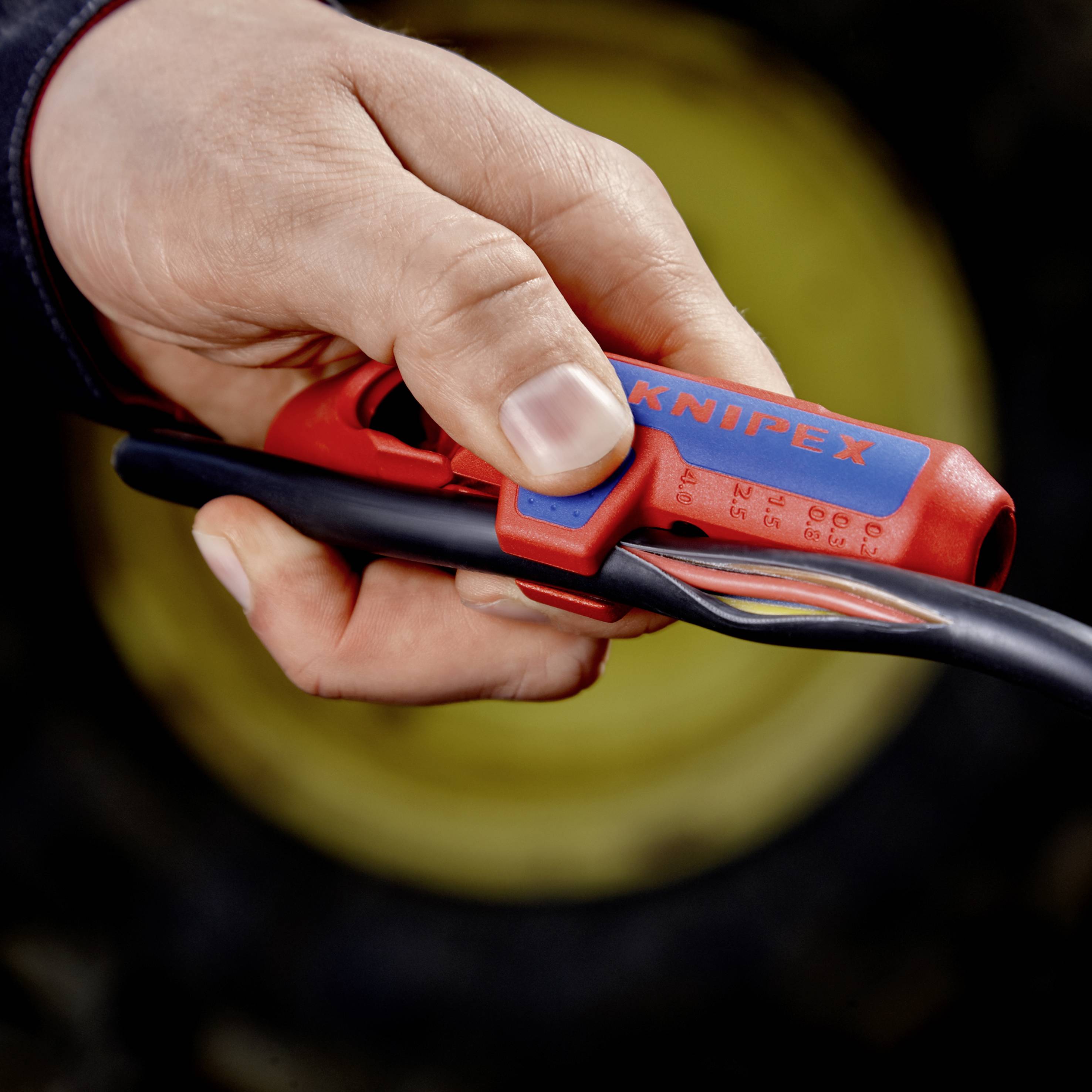 Hand hält einen roten Knipex-Kabelschneider und schneidet ein schwarzes Kabel. Fokus auf Werkzeug und Kabel, Hintergrund unscharf.
