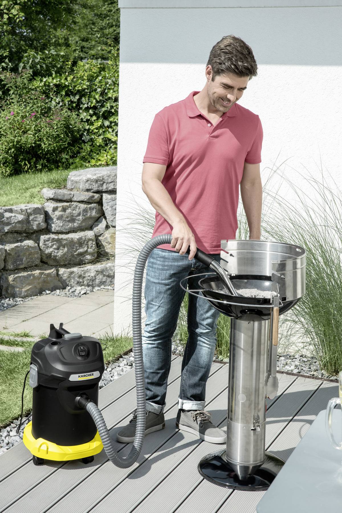Ein Mann in einem rosa Hemd saugt an einem sonnigen Tag einen Outdoor-Grill mit einem schwarz-gelben Staubsauger.