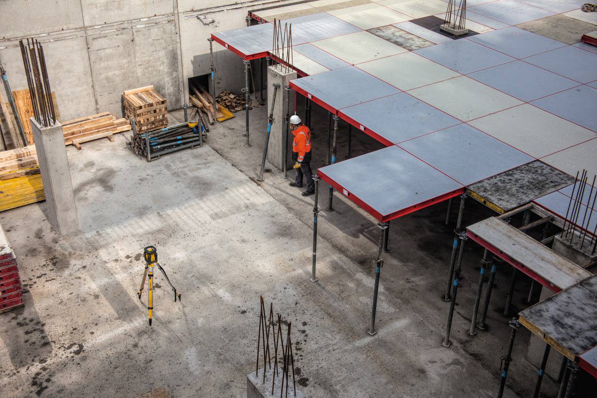Eine Baustelle mit einem Bauarbeiter in orangefarbener Weste, der unter einem teilfertigen Betongebäude steht. Posten und Stützen sichtbar.