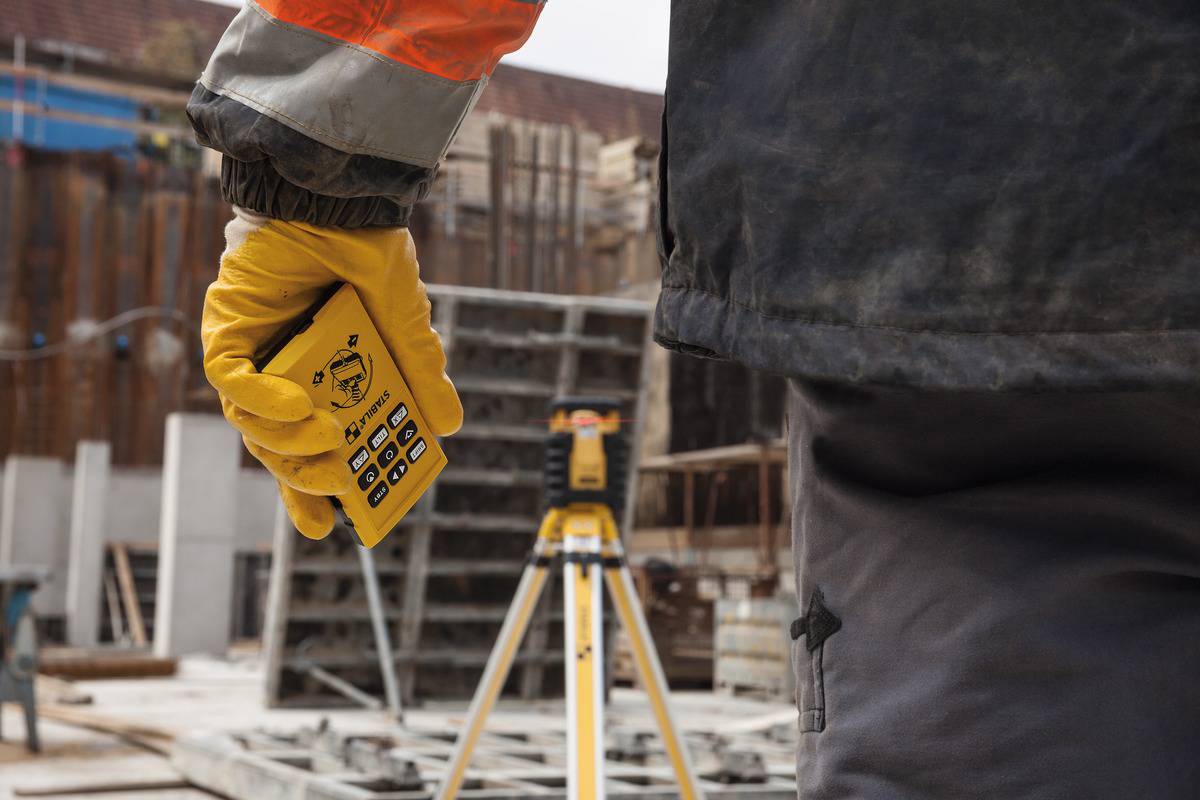 Eine Person hält ein Steuergerät und schaut auf ein orangefarbenes Vermessungsgerät auf einem Stativ auf einer Baustelle.
