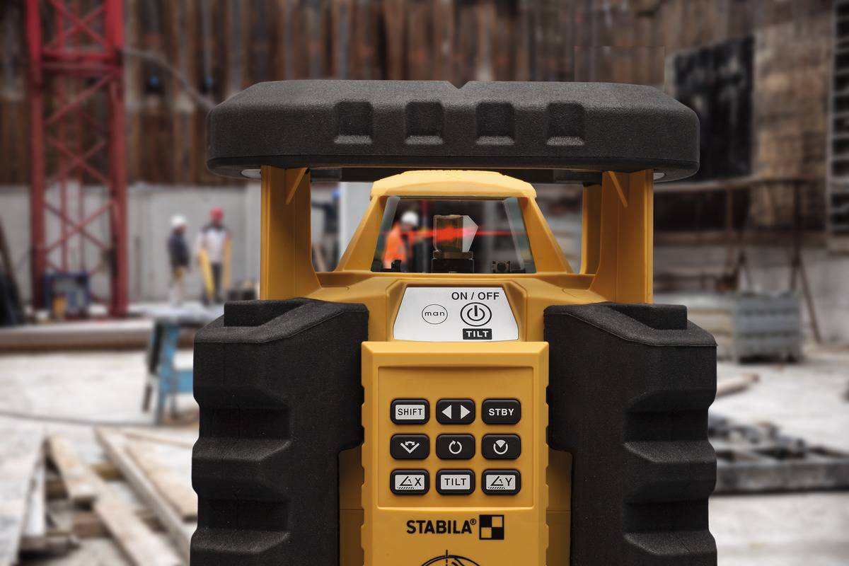 'Detailaufnahme eines gelben Lasernivelliergeräts auf einer Baustelle. Im Hintergrund unscharf arbeitende Bauarbeiter und Geräte.'