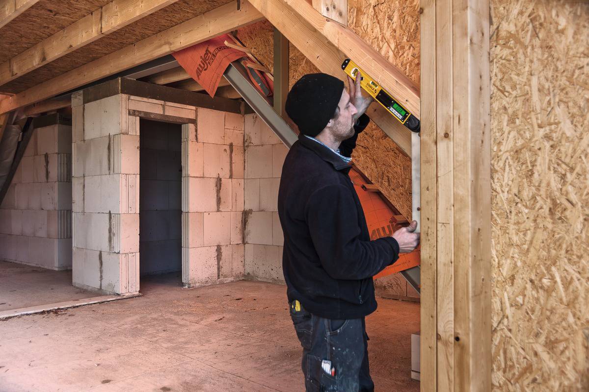 Ein Bauarbeiter überprüft mit einer Wasserwaage die vertikale Ausrichtung einer Holzwand in einem unfertigen Gebäude.