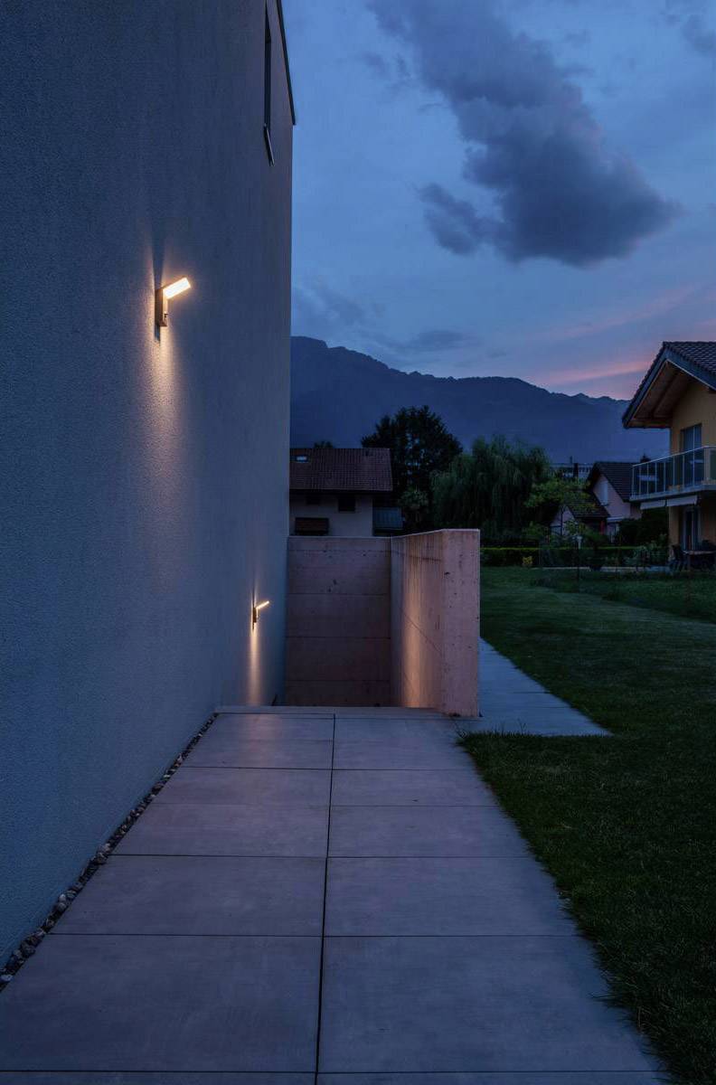 Beleuchteter Gehweg in der Dämmerung, führt hinab zu einer Treppe neben einem Haus mit Bergkulisse im Hintergrund.