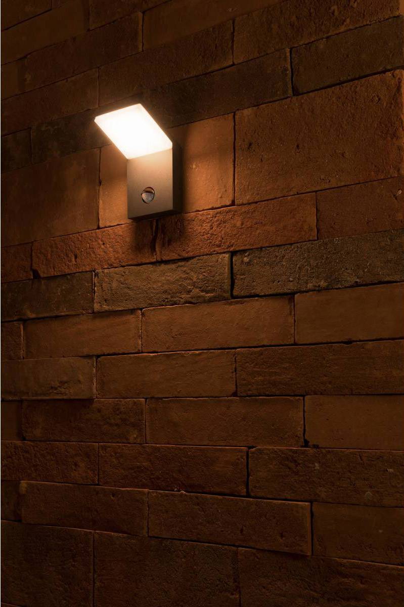 Moderne Lampe leuchtet an einer Backsteinwand und erzeugt warmes Licht. Wand hat unterschiedliche Rottöne und Texturen.