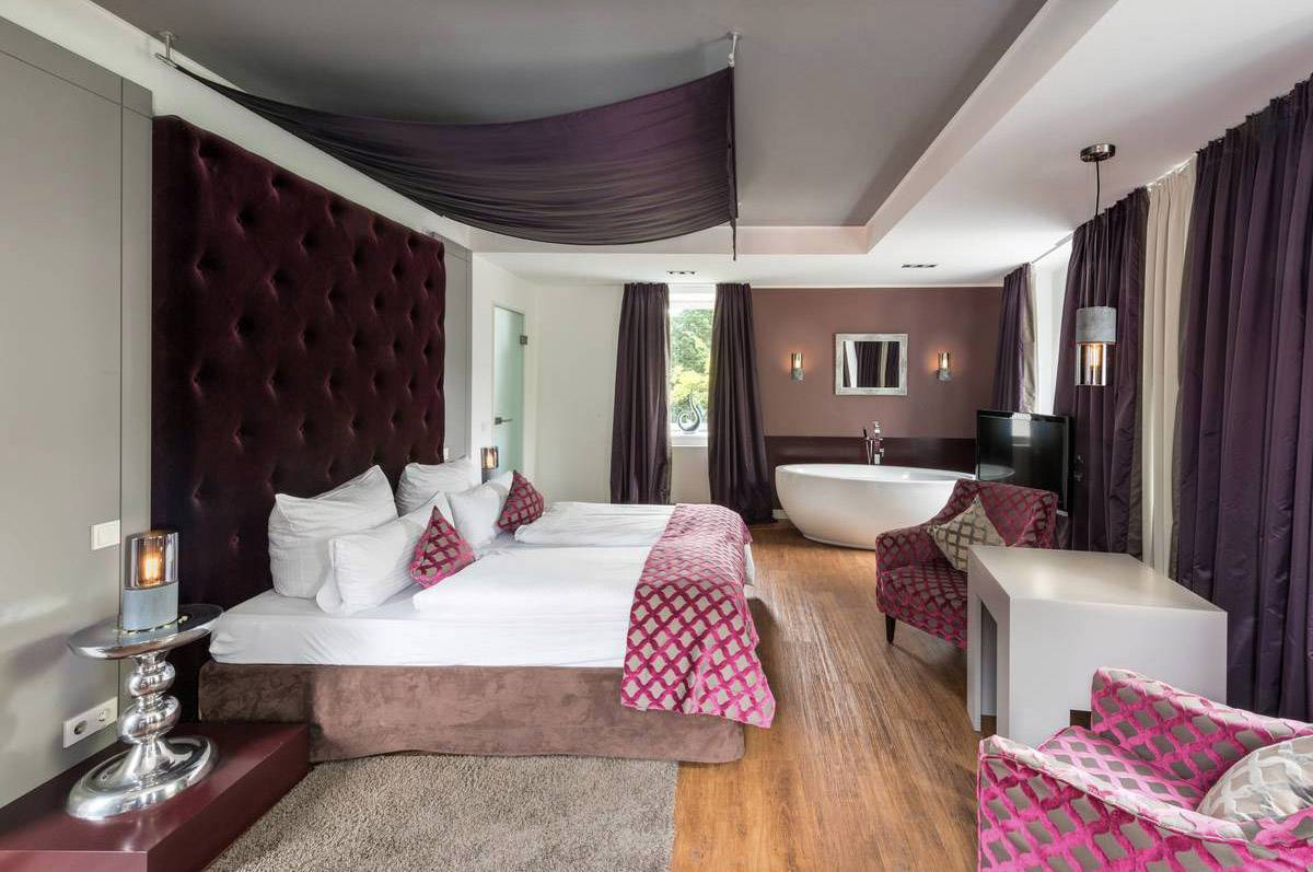 Ein luxuriöses Schlafzimmer mit großem Bett, eleganten Textilien in Rot- und Brauntönen, freistehender Badewanne und moderner Beleuchtung.