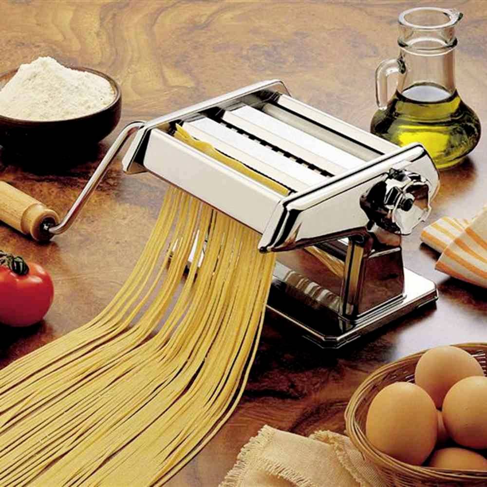 Nudelmaschine auf einem Holztisch mit frischer Pasta, Mehl, Eiern, Öl und einer Tomate daneben. Utensilien zum Pasta-Herstellen.