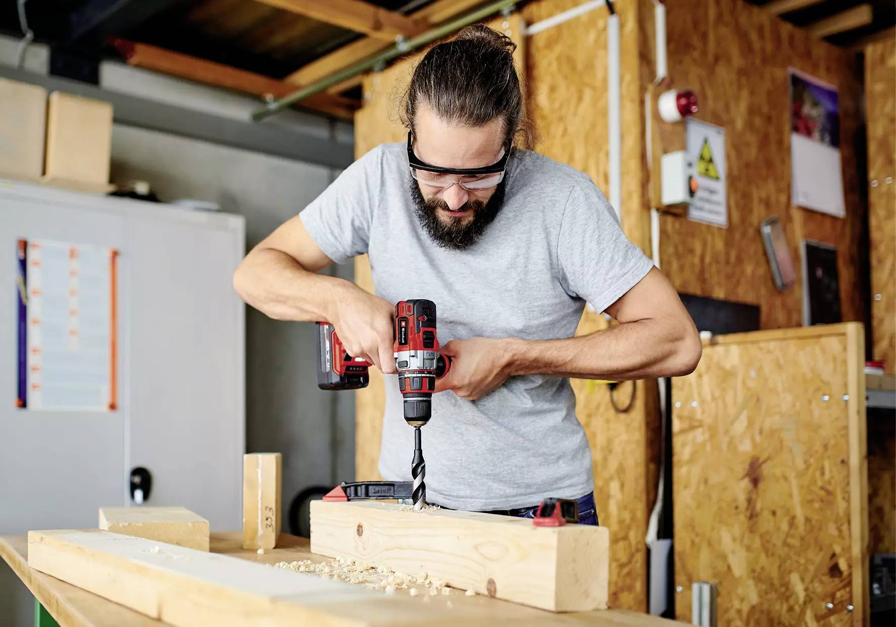 Mann mit Schutzbrille, der in einer Werkstattumgebung eine Bohrmaschine verwendet, um Löcher in Holzbretter zu bohren.