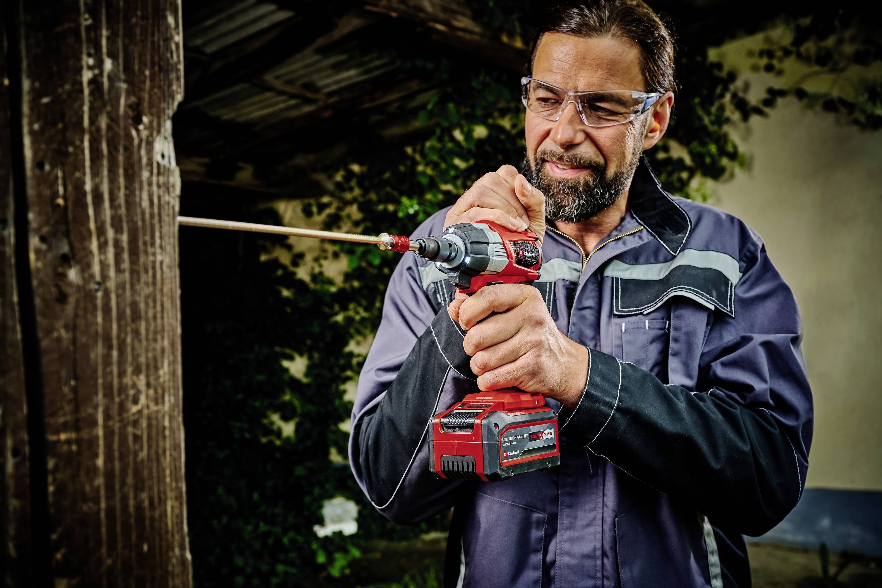 Ein Mann trägt eine Schutzbrille und benutzt eine elektrische Bohrmaschine, um ein großes Stück Holz in einer Werkstattumgebung zu bohren.