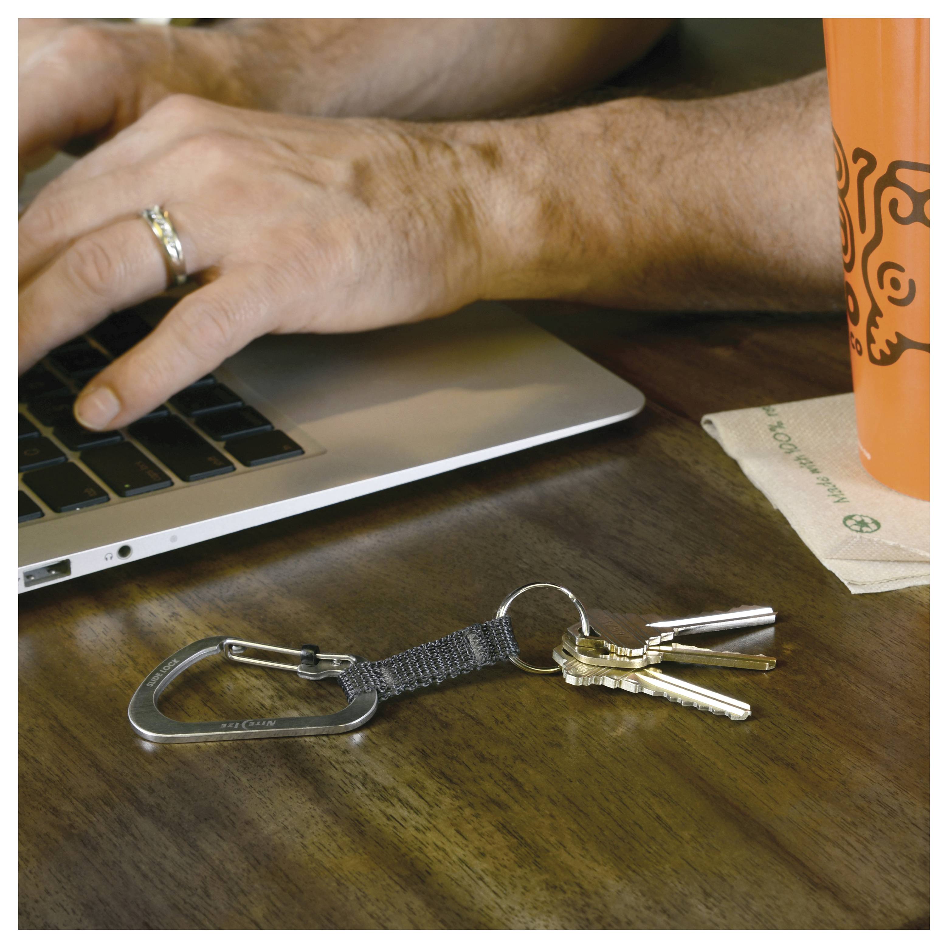 Eine Hand einer Person tippt auf einem Laptop. Auf dem Holztisch liegt ein Schlüsselanhänger mit Schlüsseln und einem Karabiner sowie eine orangefarbene Kaffeetasse.