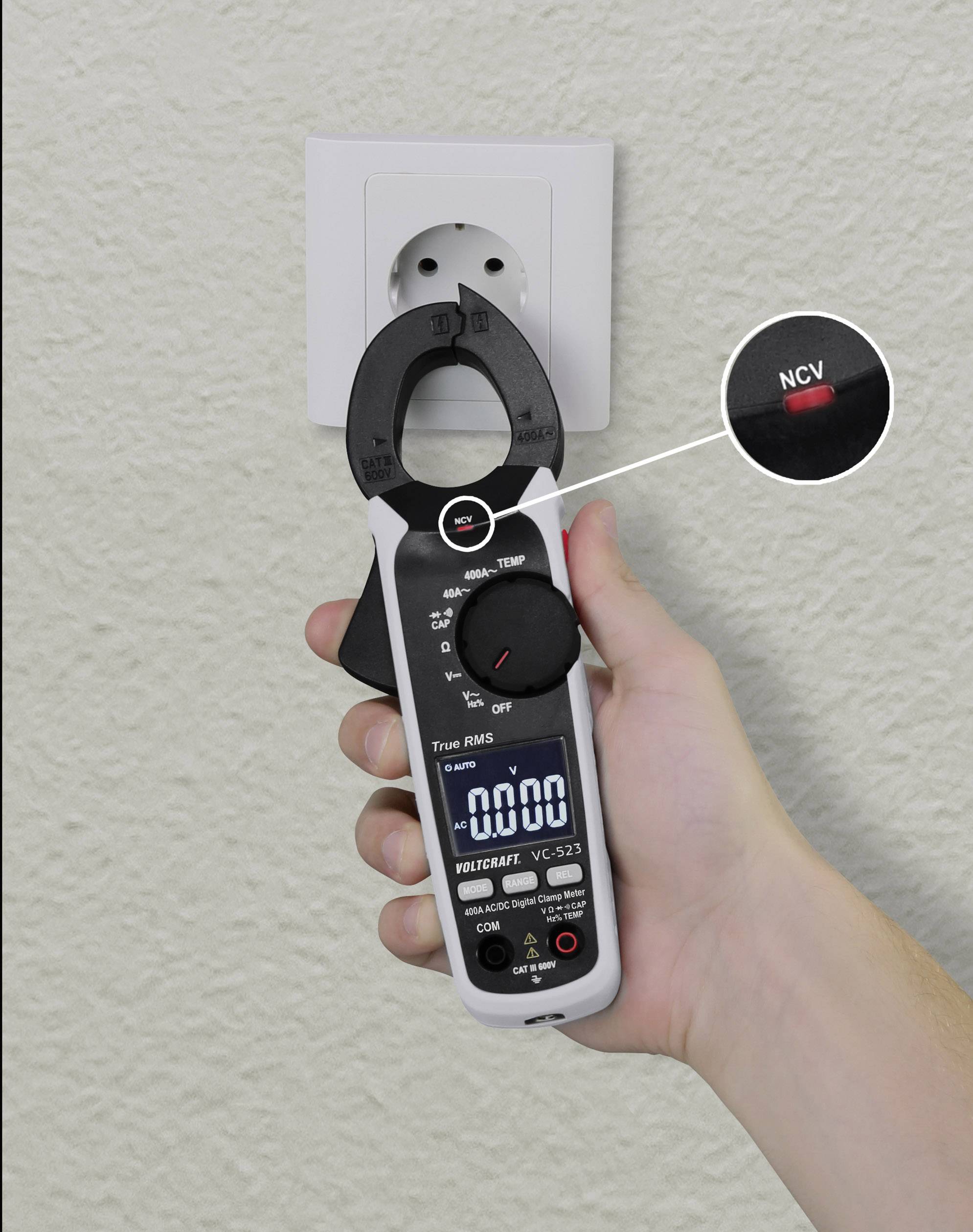 Eine Hand hält ein digitales Multimeter mit geöffneten Zangenklemmen nahe einer europäischen Steckdose. 'NCV'-Taste hervorgehoben.