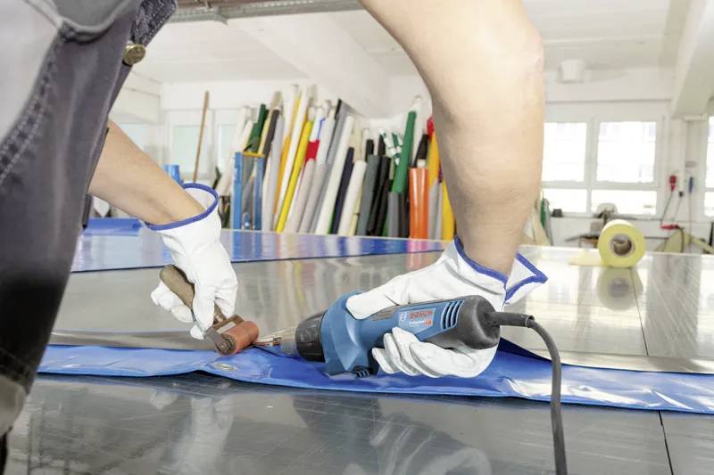 Eine Person mit Handschuhen bedient einen blauen Heißsiegelapparat an einem blauen Plastikblatt, umgeben von aufgerollten Materialien in einer Werkstattumgebung.