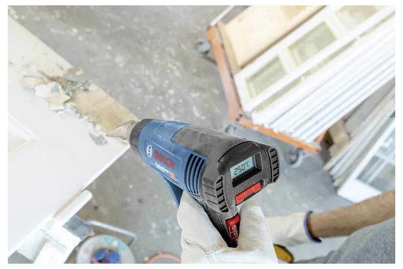 Eine Person mit Handschuhen verwendet einen Bosch-Heißluftföhn, der auf 250°C eingestellt ist, um Farbe von einer Holzoberfläche zu entfernen. Im Hintergrund sind Baumaterialien zu sehen.
