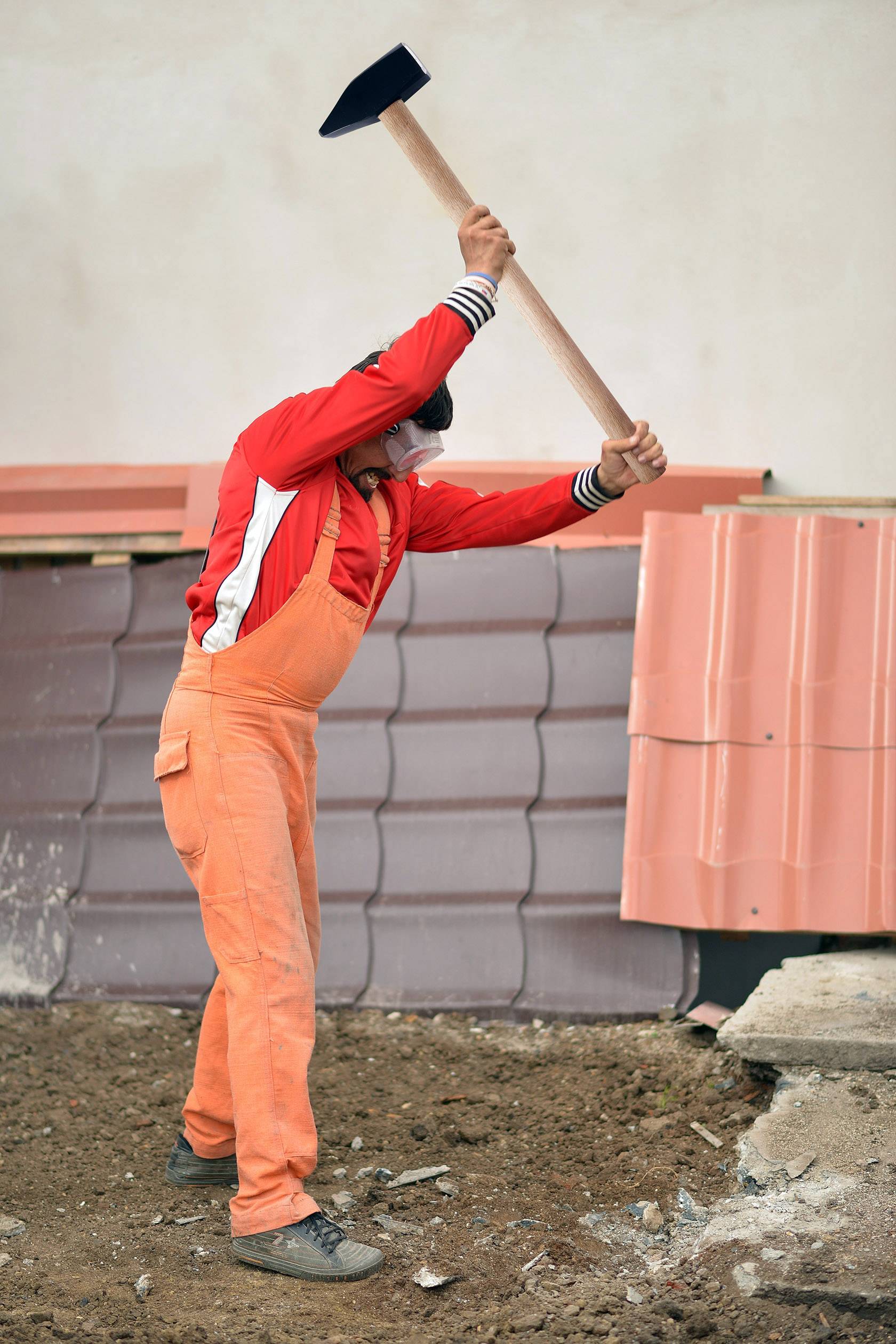 Ein Arbeiter in orangem Overall und rotem Hemd hält einen Vorschlaghammer und schlägt auf den Boden nahe einer Mauer mit Ziegeln.