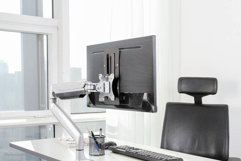 Ein Büroarbeitsplatz mit einer ergonomischen Monitorhalterung, die den Bildschirm über dem Schreibtisch schwenkbar montiert. Im Hintergrund ein Fenster mit Vorhängen.