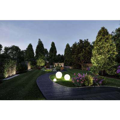 Ein gepflegter Garten bei Dämmerung mit beleuchtetem, gewundenem Weg, leuchtenden Kugellampen und bunten Blumenbeeten, umgeben von hohen Bäumen.
