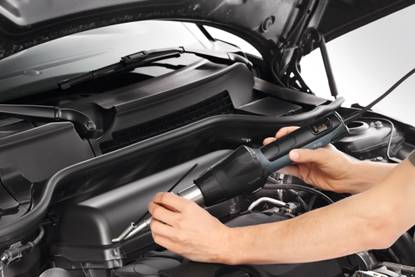 Person untersucht den Motorraum eines Autos mit einem Diagnosegerät. Der Motorhaube ist geöffnet.
