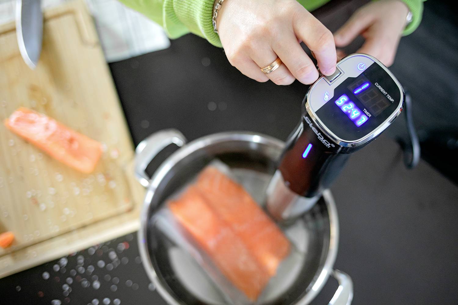 Eine Person verwendet ein Sous-Vide-Gerät, um Lachs in einem Topf zu garen. Der Timer zeigt '22:27' an. Ein Schneidebrett mit einem Stück Lachs liegt daneben.