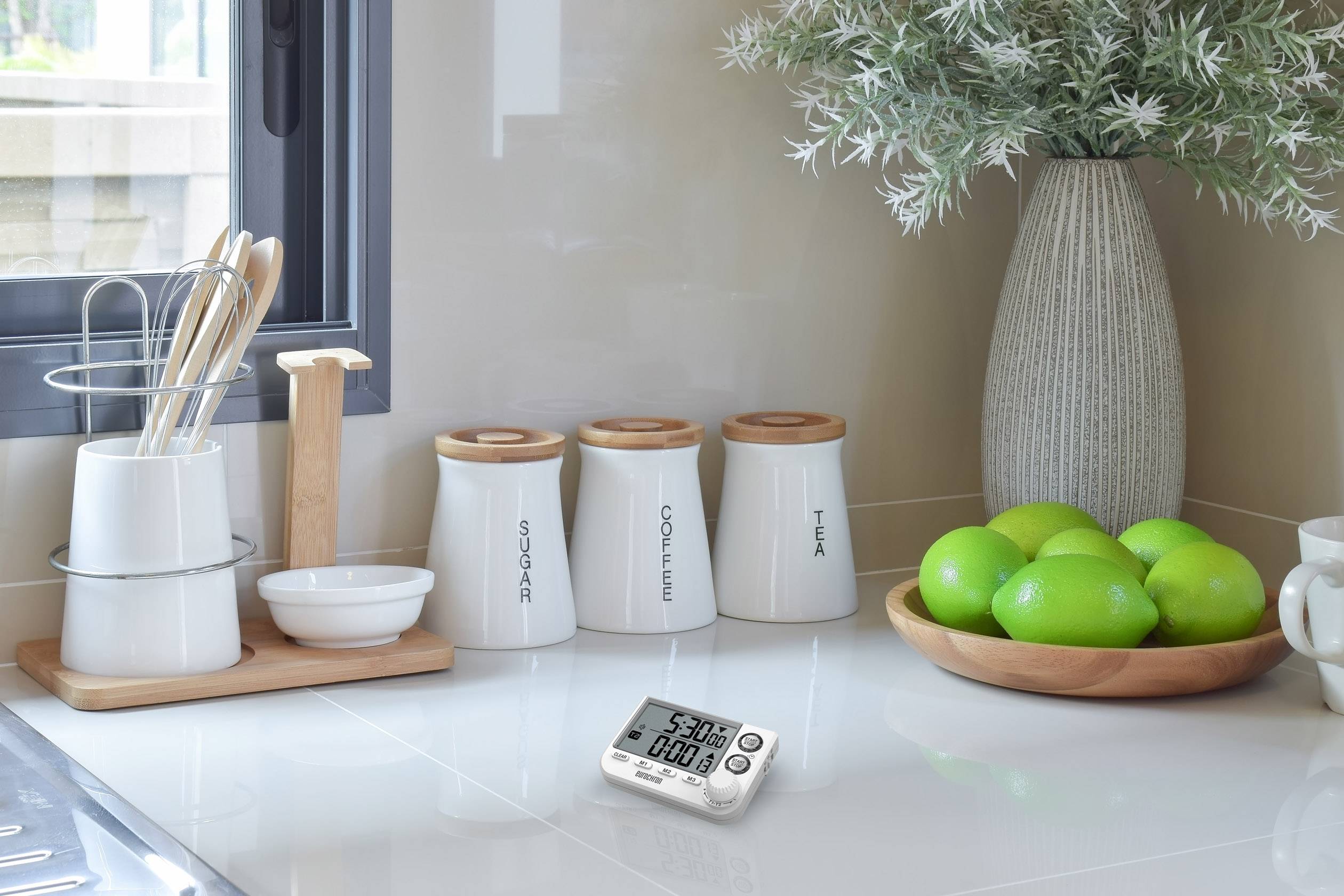 Küchentheke mit Vorratsgläsern für Zucker, Kaffee und Tee, digitalem Timer und Schale mit grünen Limetten.