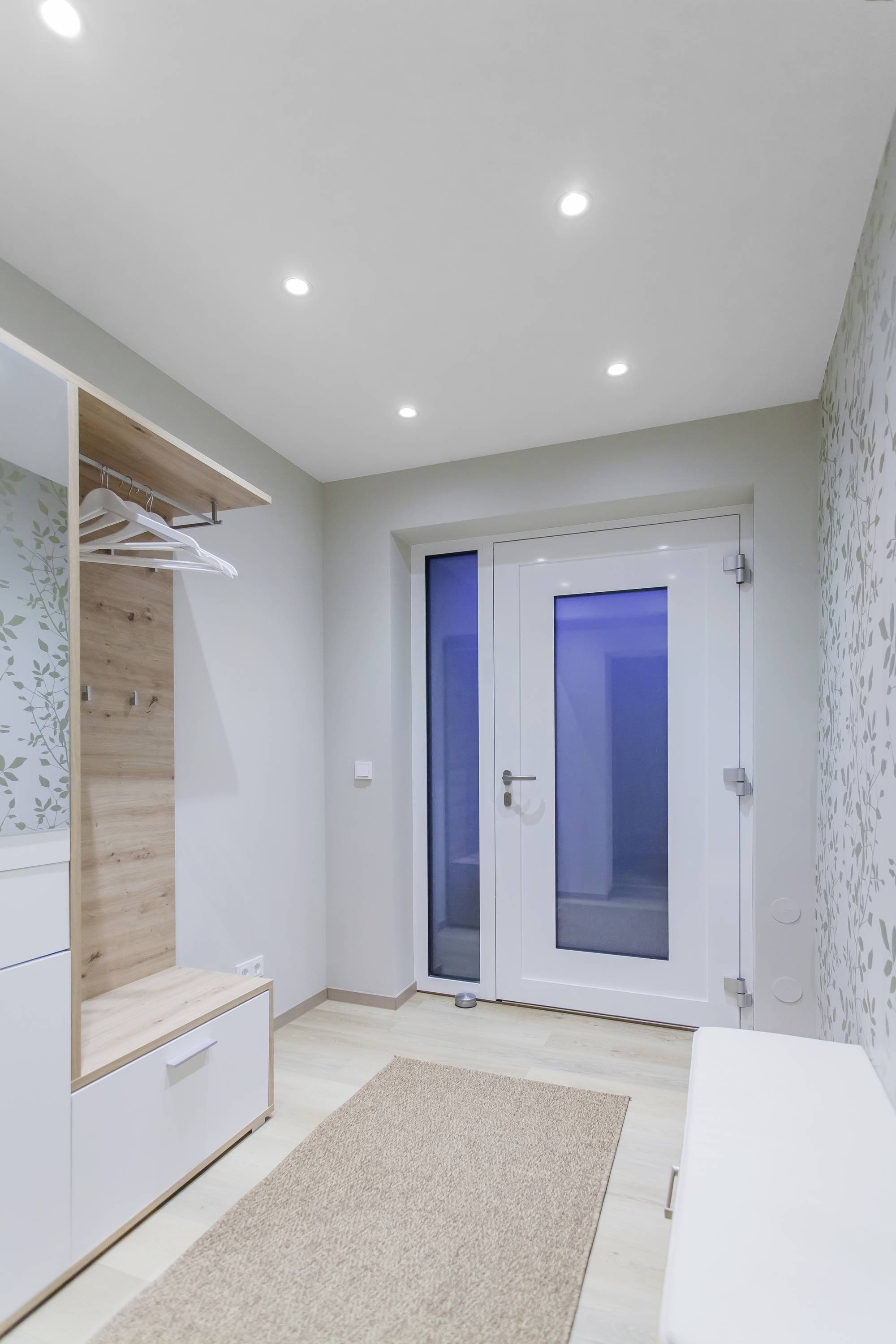 Flur mit weißer Eingangstür, kleinem Teppich, weißem Wandschrank mit Holzverkleidung und Kleiderbügeln, helle Decke mit Spotlichtern.