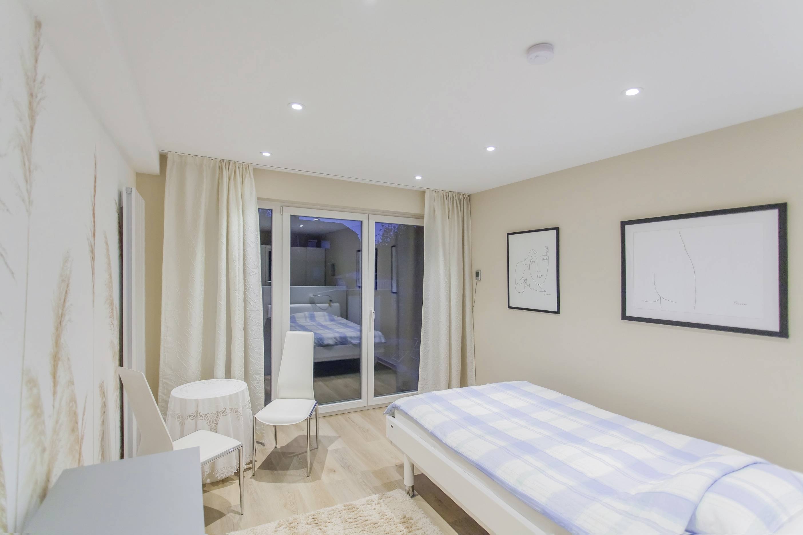 Helles Schlafzimmer mit Einzelbett, weißer Bettwäsche und einem kleinen Tisch mit zwei Stühlen. Große Fenster zeigen eine Terrasse.