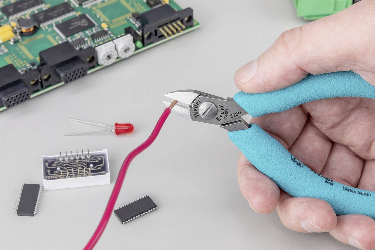 Hand hält Seitenschneider und schneidet ein rotes Kabel. Im Hintergrund liegen elektronische Bauteile und eine Platine.
