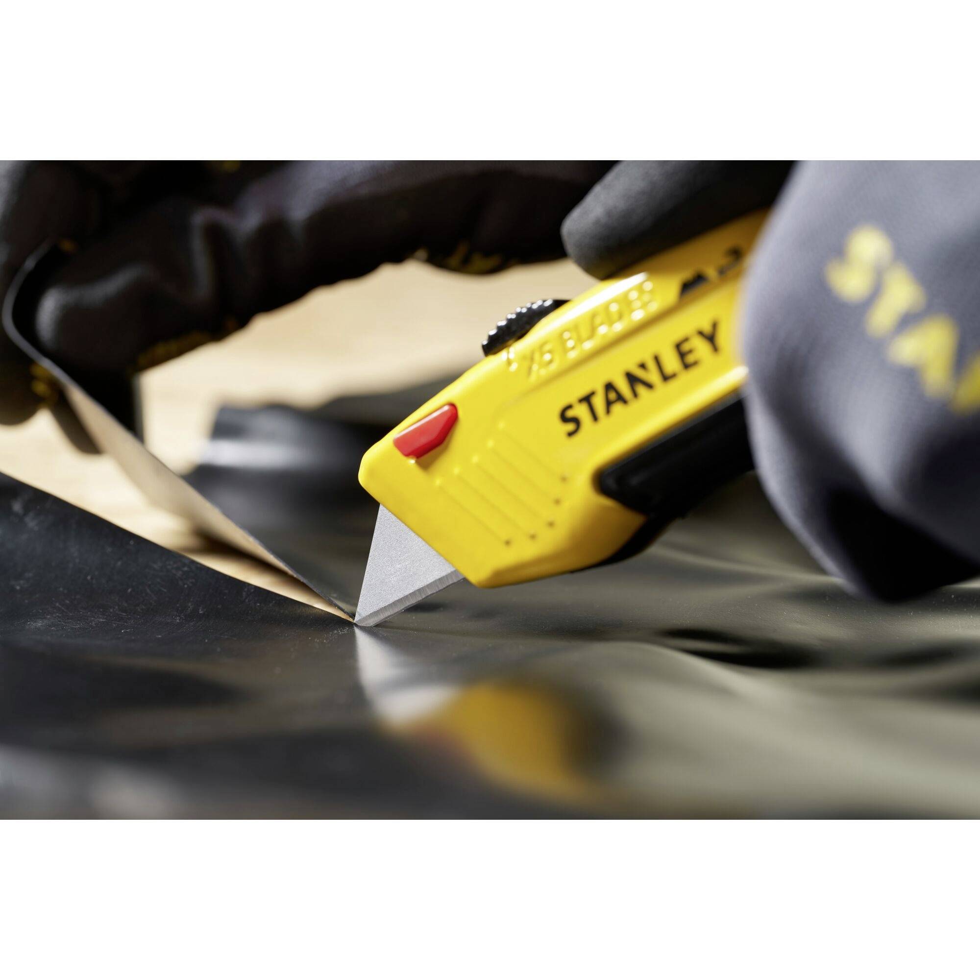 Eine Hand im Handschuh hält ein gelbes Teppichmesser der Marke Stanley, das durch ein Stück schwarzes Material schneidet.