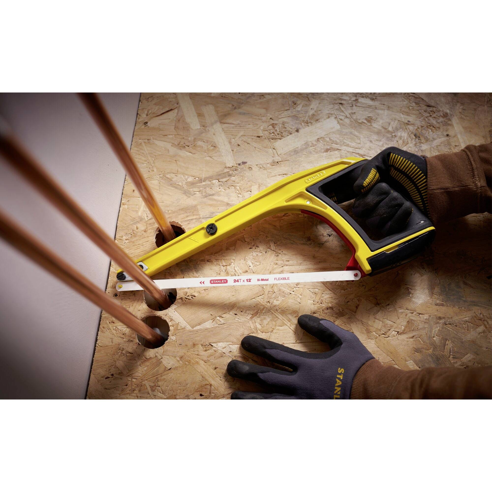 Eine Person schneidet mit einer gelben Metallsäge mehrere Kupferrohre, die aus einer Holzplatte ragen. Die Hand trägt Arbeitshandschuhe.