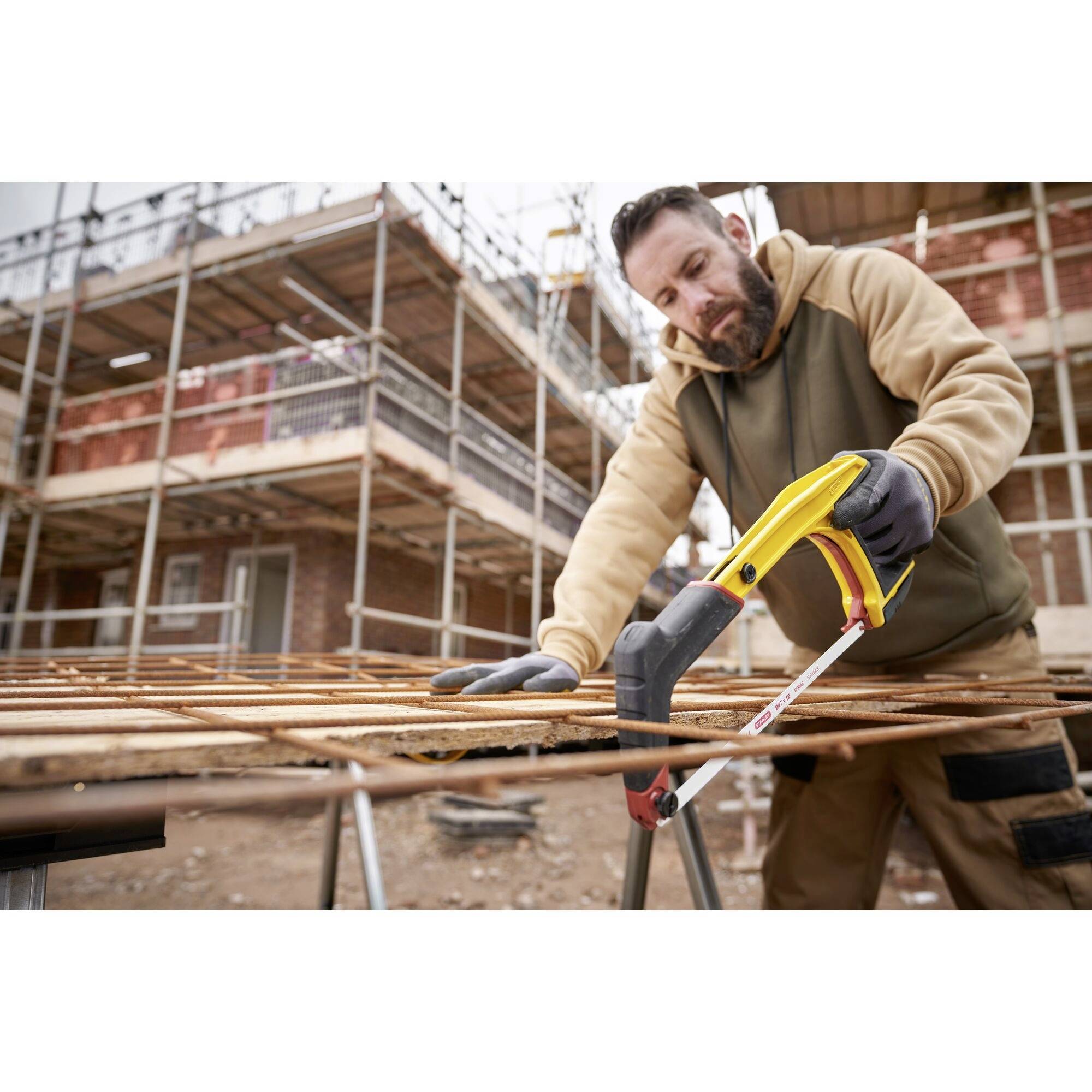 Ein Mann in Arbeitskleidung schneidet mit einer gelben Metallsäge Metallstäbe auf einer Baustelle. Hinter ihm ist ein Gebäude im Bau.