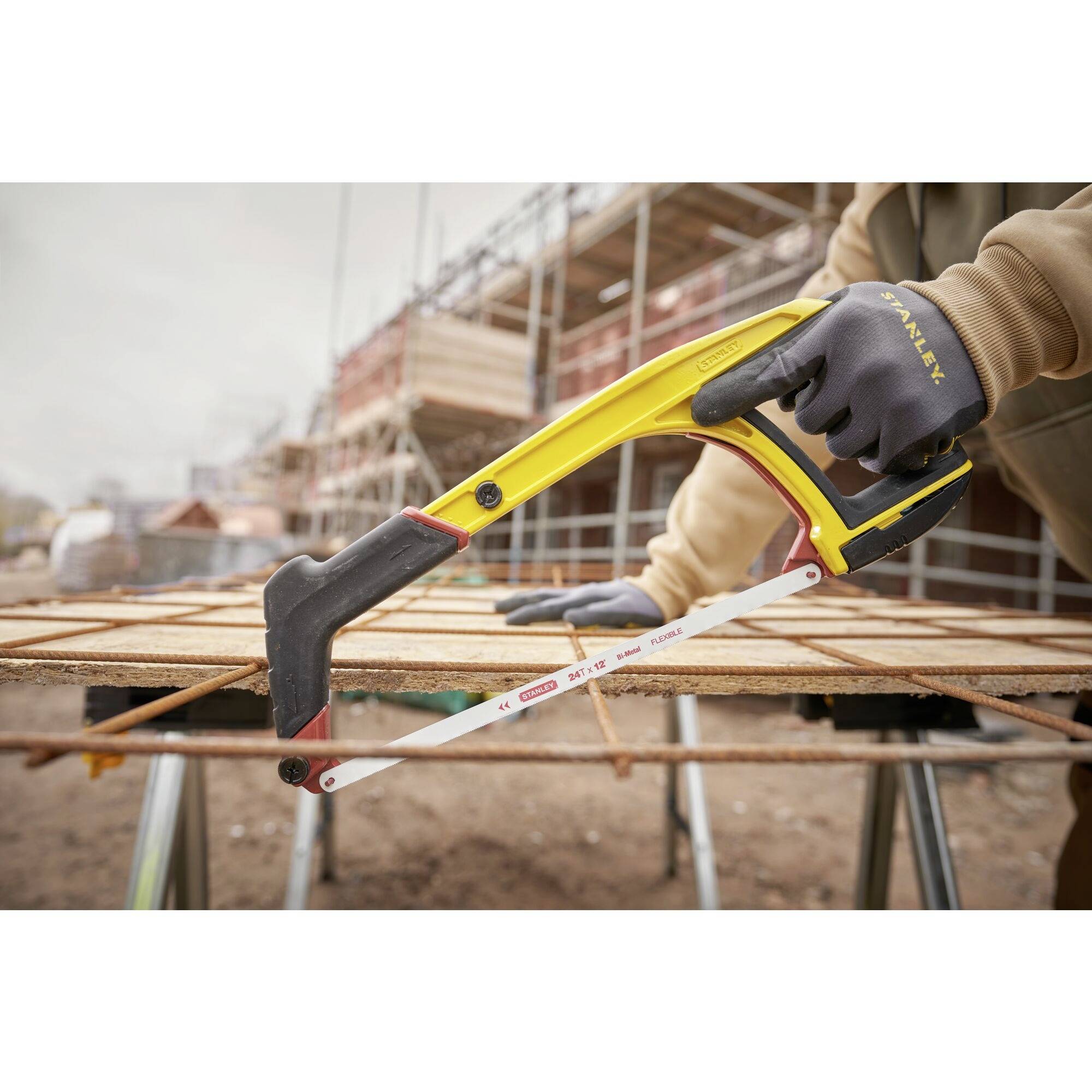 Person sägt mit Bügelsäge Metallstäbe auf Baustelle. Gebäudegerüst im Hintergrund. Fokussiert auf präzise Arbeit und Werkzeug.