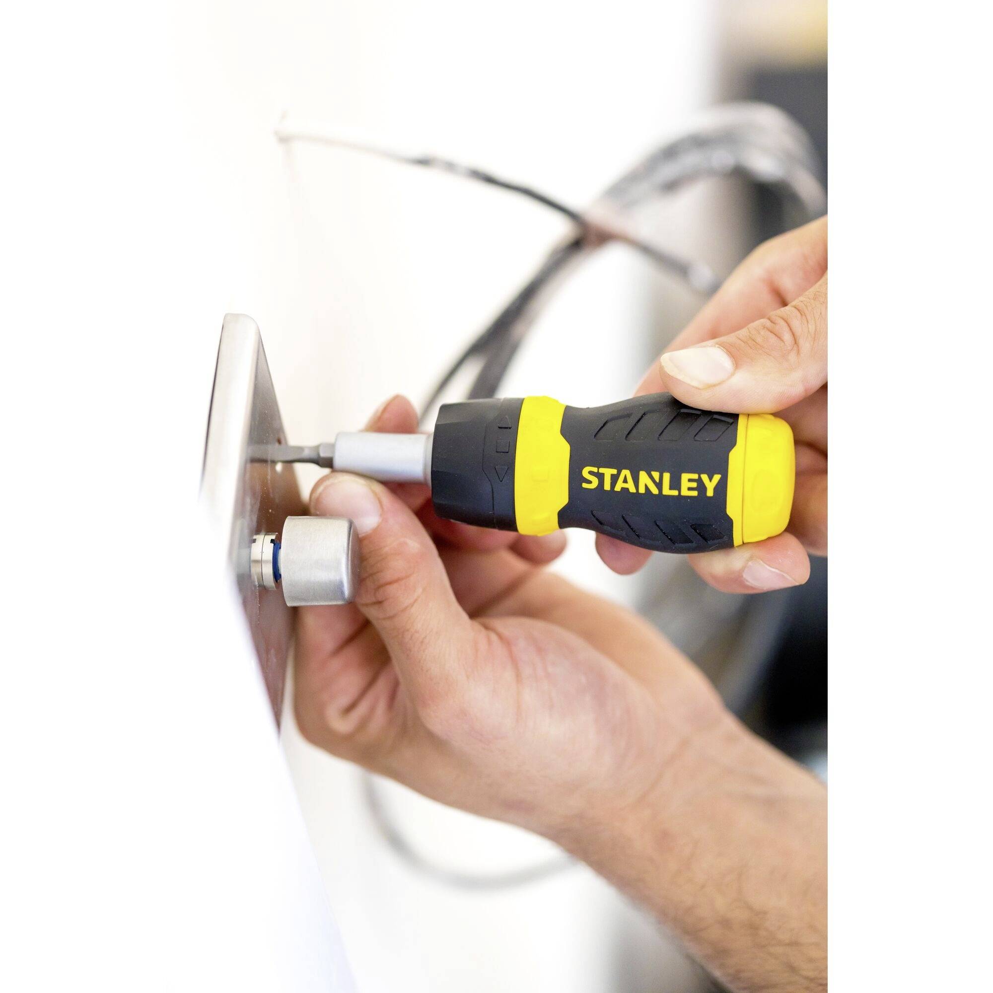 Person montiert eine Metallplatte an der Wand mit einem gelben und schwarzen Schraubendreher. Kabel im Hintergrund.