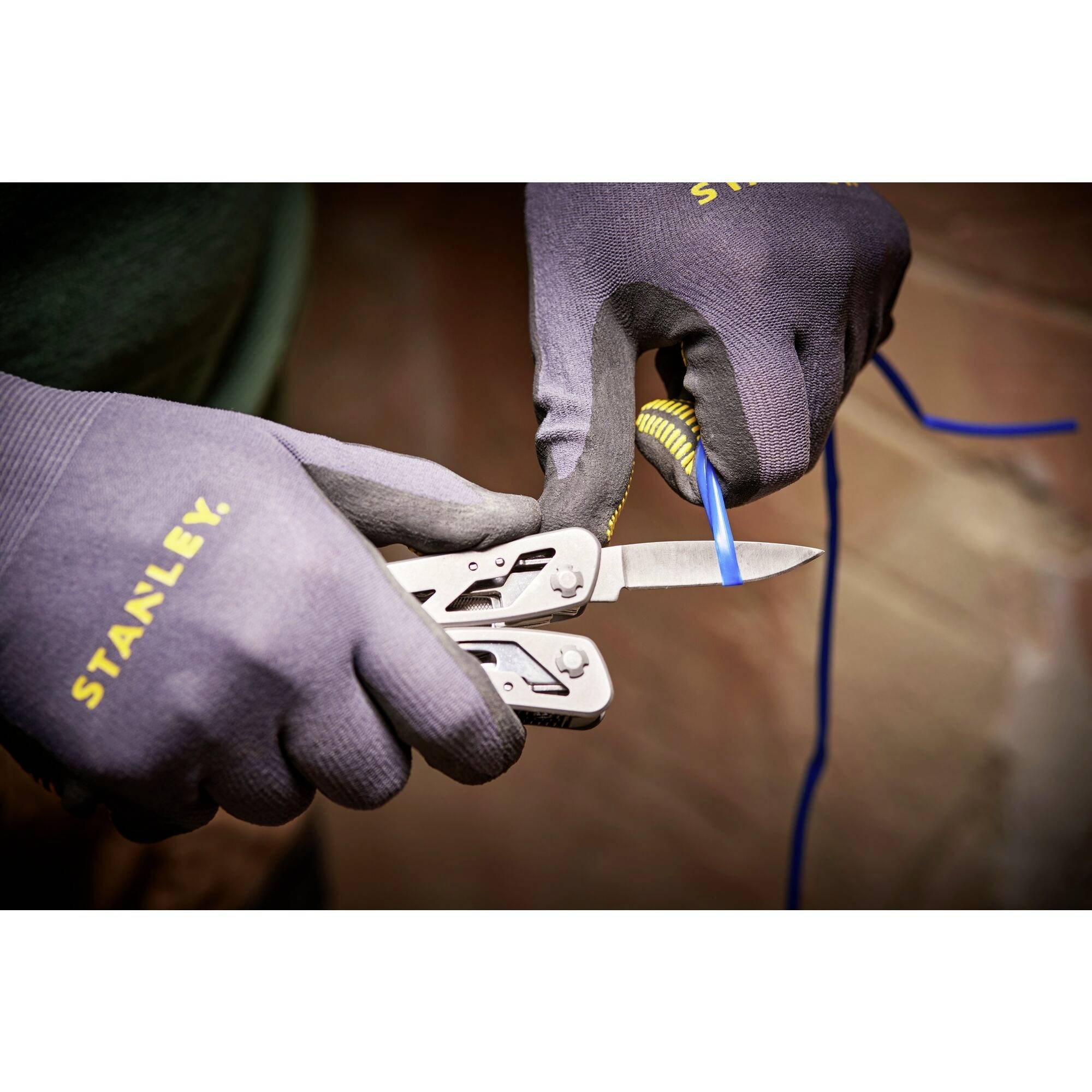 Zwei behandschuhte Hände halten ein Multifunktionswerkzeug und schneiden ein blaues Kabel. Im Hintergrund sind unscharfe Details.