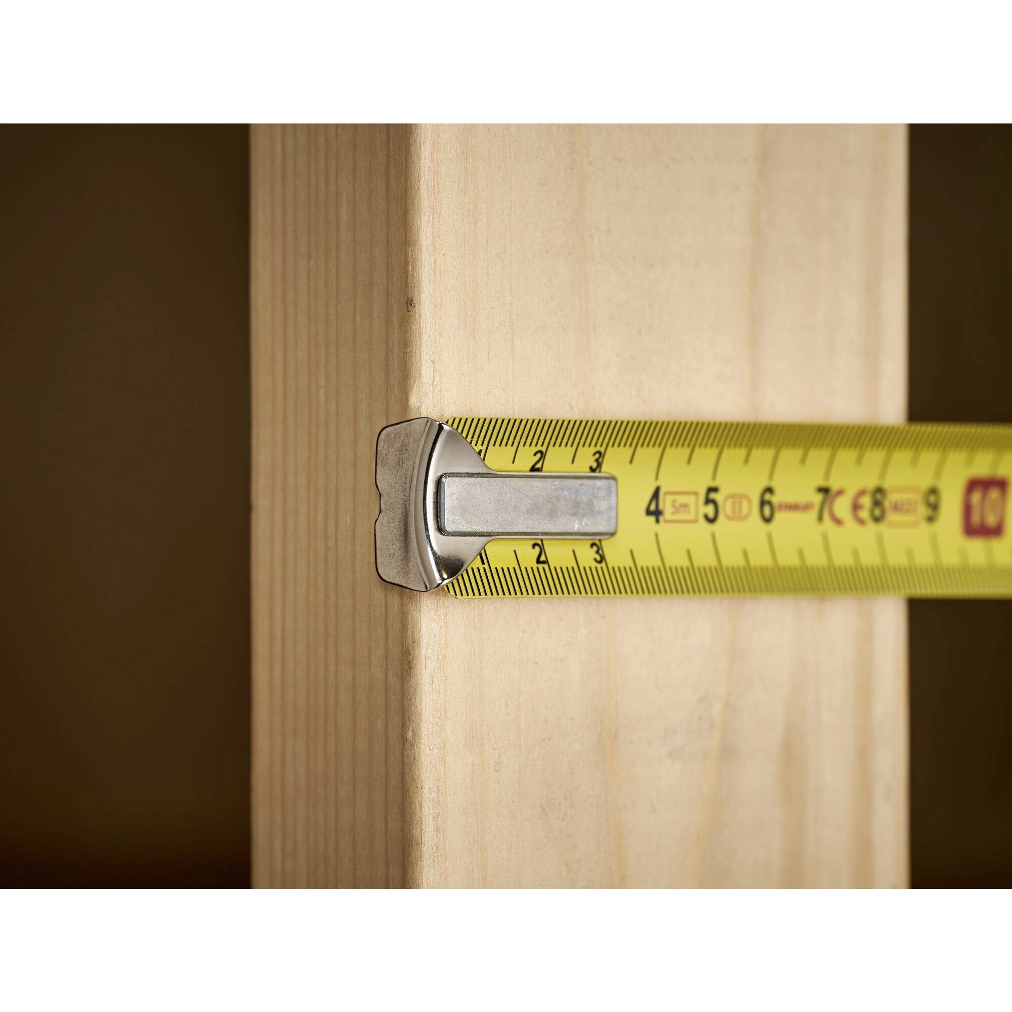 Ein gelbes Maßband misst die Breite eines Holzbretts. Das Maß beträgt 10 Zentimeter.